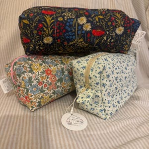 May include: Three rectangular zipped pouches made of fabric. One pouch features a dark blue background with a colourful floral pattern. Another has a cream background with dense multi-coloured flowers. The third is light blue with small blue flowers.