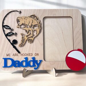 May include: A light-colored wooden picture frame featuring a cutout of a fish, a black fishing rod with two lures labeled Adilynn and Charlie, a red and white bobber, and the blue text "WE ARE HOOKED ON Daddy" engraved on the front surface.