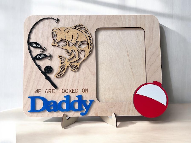 May include: A light-colored wooden picture frame featuring a cutout of a fish, a black fishing rod with two lures labeled Adilynn and Charlie, a red and white bobber, and the blue text "WE ARE HOOKED ON Daddy" engraved on the front surface.