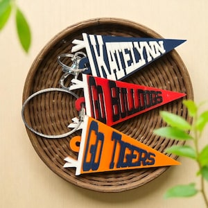May include: Three pennant-shaped keychains in a woven basket. One is navy blue with "KATELYNN" in white, another is red with "GO BULLDOGS" in black, and the last is orange with "GO TIGERS" in navy blue. Each has a silver keyring.