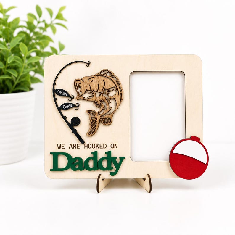 May include: A rectangular light-colored wooden picture frame featuring a laser-cut fish and a fishing rod with two small black lures labeled Annalee and Charlie. The text reads WE ARE HOOKED ON Daddy in green, with a red and white fishing bobber attached.