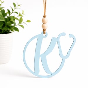 May include: A light blue hanging ornament shaped like the letter K integrated with a stethoscope silhouette. The ornament is suspended by a tan twine cord featuring two smooth, round, light-colored wooden beads above the letter.