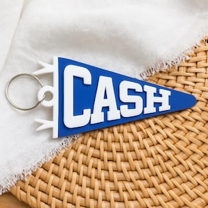 May include: A blue pennant-shaped keychain with the word "CASH" in white block letters. The keychain has a silver ring and white accents. The pennant is resting on a woven wicker surface.