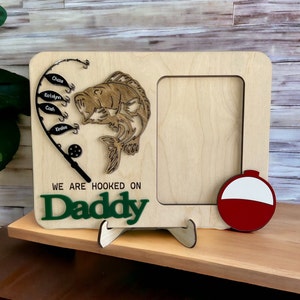 May include: A light-colored wooden picture frame featuring a laser-cut fishing rod with four fish-shaped tags labeled Chase, Katelyn, Cash, and Kinslee. A wooden fish, a red and white bobber, and the text WE ARE HOOKED ON Daddy are attached.