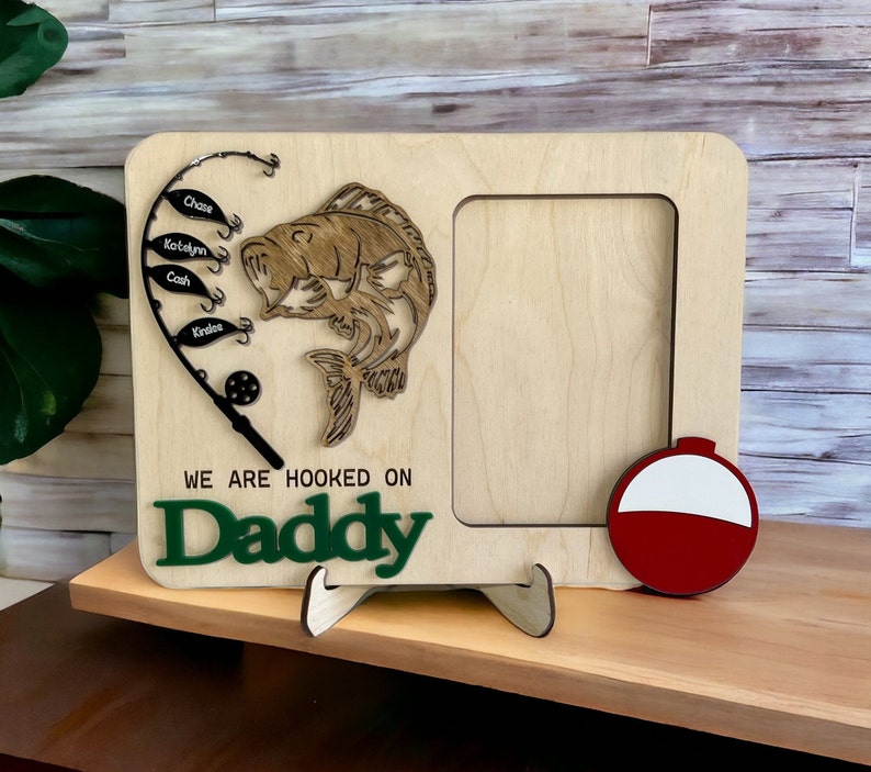 May include: A light-colored wooden picture frame featuring a laser-cut fishing rod with four fish-shaped tags labeled Chase, Katelyn, Cash, and Kinslee. A wooden fish, a red and white bobber, and the text WE ARE HOOKED ON Daddy are attached.