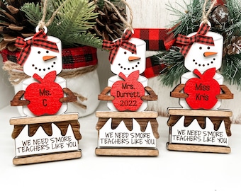 Puede incluir: Tres adornos de madera de muñeco de nieve con detalles de manzana roja y el texto "We need s'more teachers like you". Los muñecos de nieve sostienen manzanas con los nombres "Ms. C", "Mrs. Durrett 2022" y "Miss Kris" escritos en ellas.