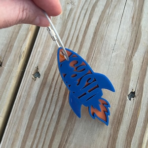 May include: A blue rocket-shaped keychain with the word "SHISH" cut out. The rocket has orange accents and a silver wire loop for attachment. The keychain is held by a hand against a wooden background.