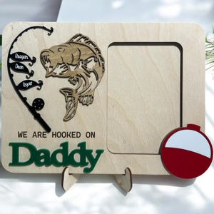 May include: A rectangular light wood picture frame featuring a laser-cut black fishing rod with three fish-shaped tags labeled Reagan, Dean, and Ryver, a wooden fish cutout, a red and white bobber, and the text WE ARE HOOKED ON Daddy in green.
