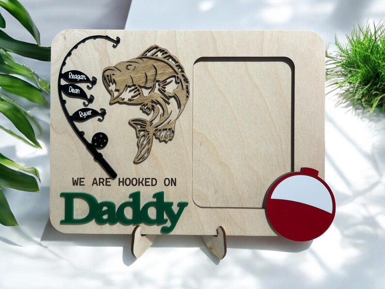 May include: A rectangular light wood picture frame featuring a laser-cut black fishing rod with three fish-shaped tags labeled Reagan, Dean, and Ryver, a wooden fish cutout, a red and white bobber, and the text WE ARE HOOKED ON Daddy in green.