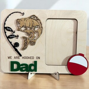 May include: A light-colored wooden picture frame featuring a cutout of a fish, a fishing rod with two lures labeled Darby and Tristan, and a red and white fishing bobber. The text WE ARE HOOKED ON Dad is engraved and attached in green lettering.