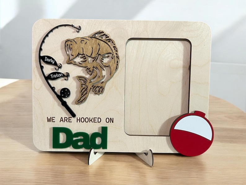 May include: A light-colored wooden picture frame featuring a cutout of a fish, a fishing rod with two lures labeled Darby and Tristan, and a red and white fishing bobber. The text WE ARE HOOKED ON Dad is engraved and attached in green lettering.
