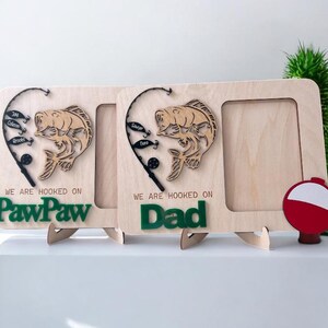 May include: Two light-colored wooden picture frames featuring a laser-cut fishing rod, a fish, and small lures. The text reads "WE ARE HOOKED ON" with "PawPaw" or "Dad" in green. A red and white fishing bobber is attached to the right frame.