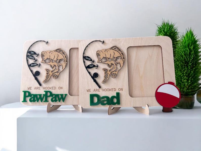 May include: Two light-colored wooden picture frames featuring a laser-cut fishing rod, a fish, and small lures. The text reads "WE ARE HOOKED ON" with "PawPaw" or "Dad" in green. A red and white fishing bobber is attached to the right frame.
