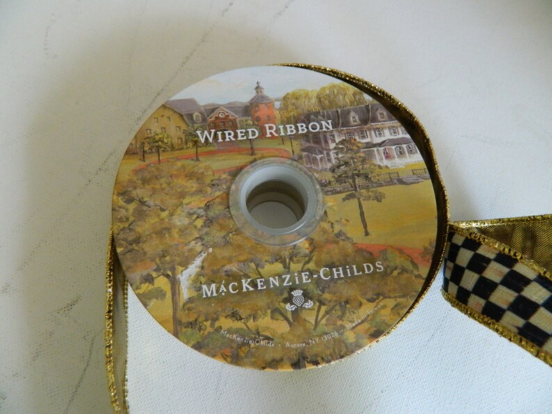May include: A roll of gold wired ribbon with a black and white checkered ribbon. The ribbon features a printed image of a house and the text "Wired Ribbon" and "MACKENZIE-CHILDS".