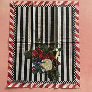 Puede incluir: Una servilleta de tela a rayas blancas y negras con un borde de bast&oacute;n de caramelo rojo y blanco. El centro de la servilleta presenta un dise&ntilde;o floral con bayas rojas, flores blancas y agujas de pino.