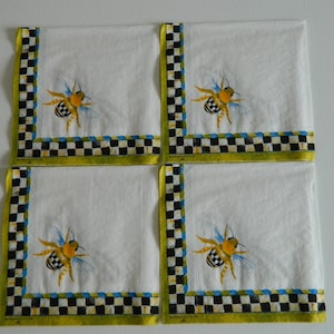 May include: Set of four white linen napkins with a black and white chequered border and a yellow, black and blue bee illustration. The napkins have a green border with the text "McKenzie-Childs" printed on the bottom.
