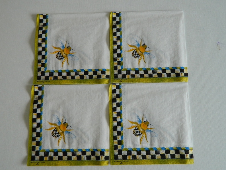 May include: Set of four white linen napkins with a black and white chequered border and a yellow, black and blue bee illustration. The napkins have a green border with the text "McKenzie-Childs" printed on the bottom.