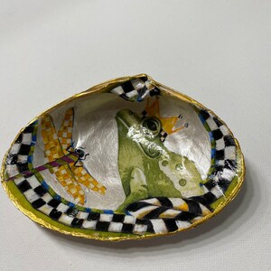 May include: A decorative seashell dish with a black and white checkered pattern and gold trim. The dish features a painted image of a green frog wearing a crown and a dragonfly.