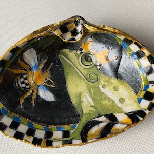 May include: A hand-painted seashell dish with a black and white checkered border. The dish features a green frog with a gold crown, a bee with black and white stripes, and gold accents.