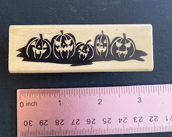 May include: A wooden stamp with a black silhouette of five jack-o'-lantern pumpkins with carved faces. The pumpkins are arranged in a row and are all facing the viewer.