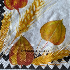 May include: White paper napkin with a black and white chequered border. The napkin features a floral design with yellow and red leaves and wheat stalks. The text "MACKENZIE-CHILDS" is printed in black on the napkin.