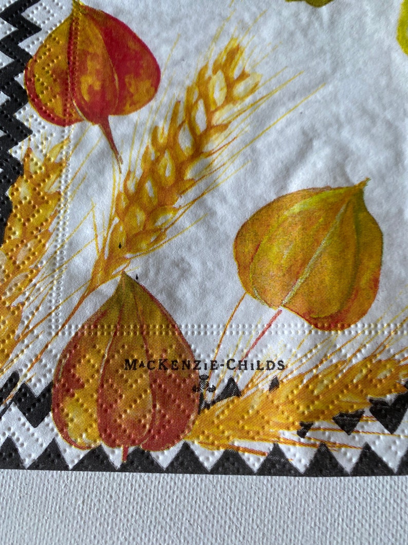 May include: White paper napkin with a black and white chequered border. The napkin features a floral design with yellow and red leaves and wheat stalks. The text "MACKENZIE-CHILDS" is printed in black on the napkin.