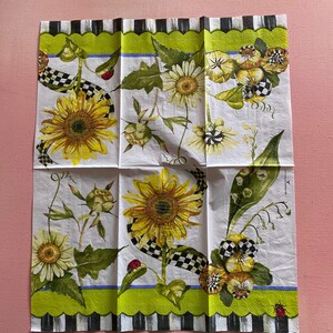 Puede incluir: Una servilleta de papel blanca con un borde a cuadros verde, negro y blanco. La servilleta presenta un dise&ntilde;o floral con girasoles, margaritas y pensamientos. Tambi&eacute;n hay mariquitas y una cinta a cuadros.