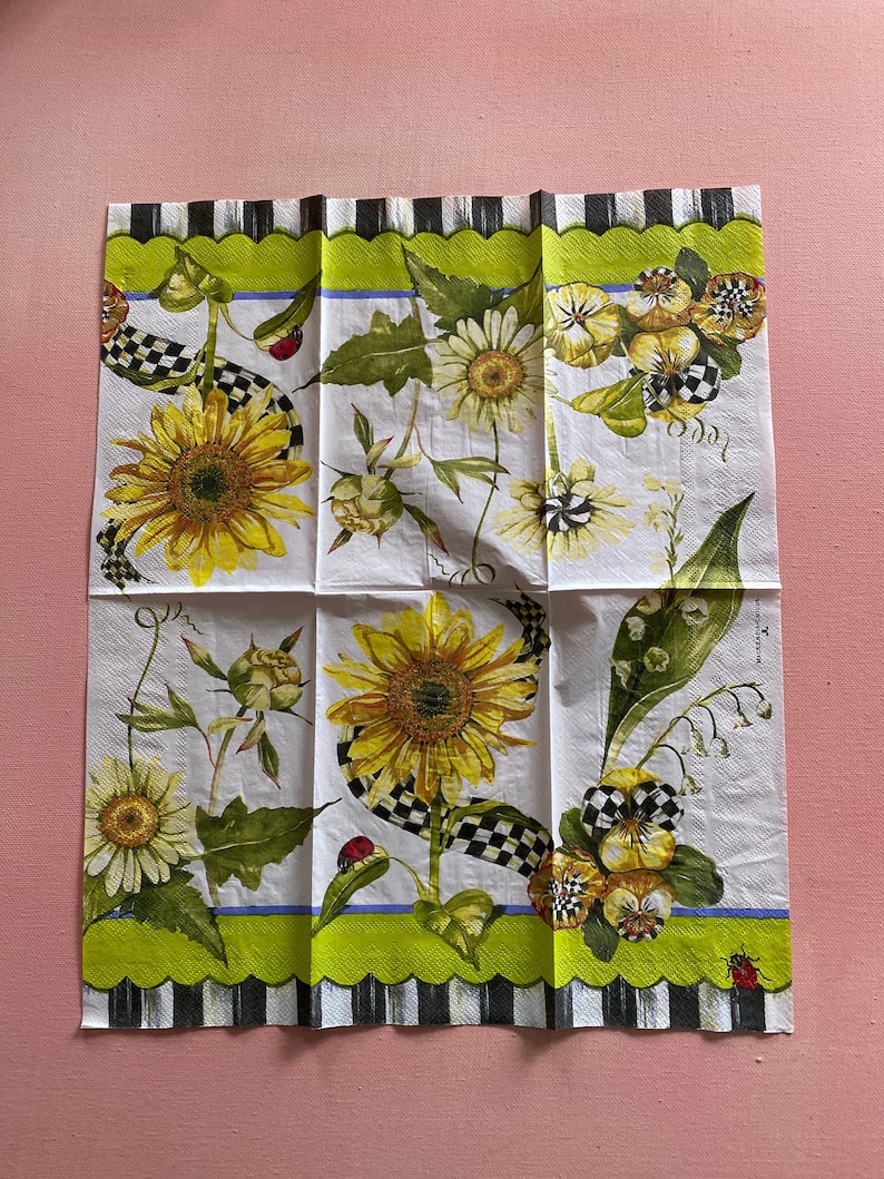 Puede incluir: Una servilleta de papel blanca con un borde a cuadros verde, negro y blanco. La servilleta presenta un dise&ntilde;o floral con girasoles, margaritas y pensamientos. Tambi&eacute;n hay mariquitas y una cinta a cuadros.