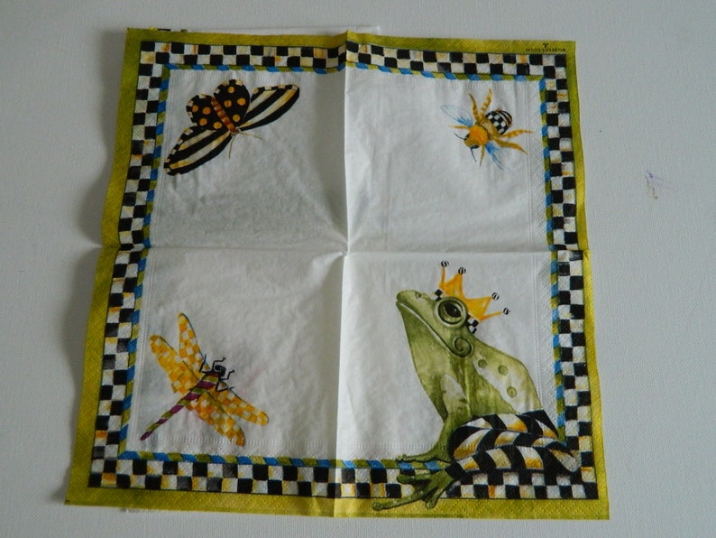 May include: A white paper napkin featuring a green frog wearing a gold crown, a yellow dragonfly, a black and yellow butterfly, and a yellow bee. The napkin has a checkered black and white border with a yellow background.
