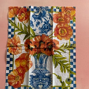 Puede incluir: Una servilleta blanca con un dise&ntilde;o floral azul y blanco. El dise&ntilde;o presenta dos jarrones con motivos florales y amapolas rojas. El borde de la servilleta es un patr&oacute;n de cuadros azules y blancos.