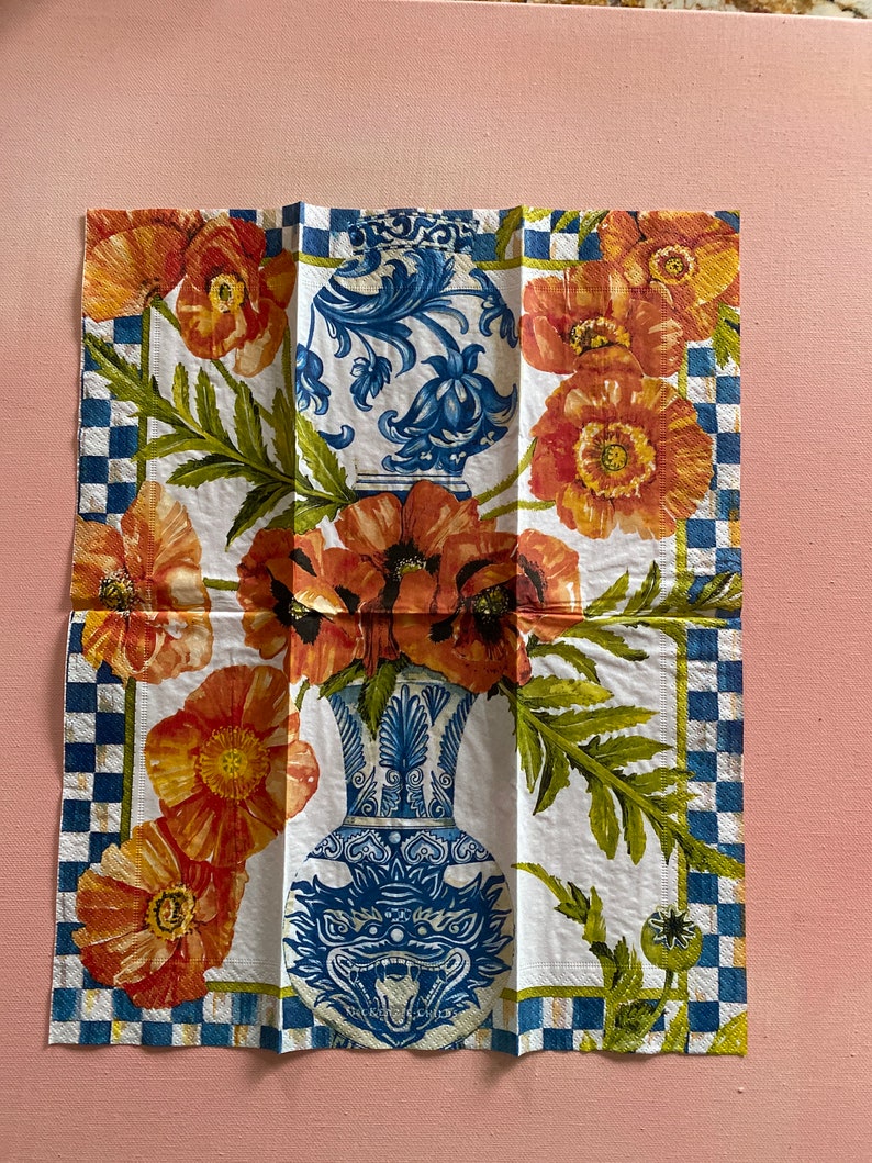 Puede incluir: Una servilleta blanca con un dise&ntilde;o floral azul y blanco. El dise&ntilde;o presenta dos jarrones con motivos florales y amapolas rojas. El borde de la servilleta es un patr&oacute;n de cuadros azules y blancos.
