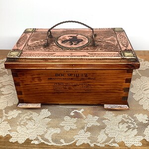 May include: A vintage wooden box with a copper-coloured lid and handle. The lid features embossed designs and a central medallion with a silhouette. The box has text that reads "DOC SCHULZ AMAZING SNAKE OIL MEDICINE".