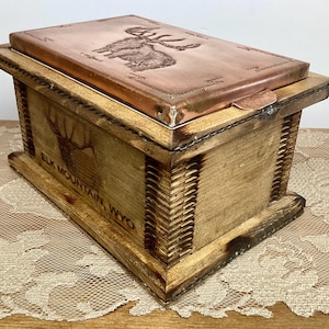 May include: Wooden box with a copper lid featuring an embossed elk design. The box is rectangular with a hinged lid and the words "ELK MOUNTAIN, WYO" are printed on the front. The box has a rustic, handcrafted appearance.