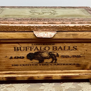 May include: A wooden box with a copper-colored lid and accents. The box is engraved with the words "Buffalo Balls" and a bison image. The lid has a textured surface with a lighter-colored center. The box is rectangular and has a hinged lid.
