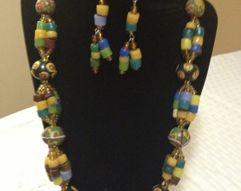 May include: A colourful beaded necklace with a red glass bead at the centre. The necklace is made of small, round beads in shades of yellow, blue, green, brown, and black. The necklace is paired with matching earrings.