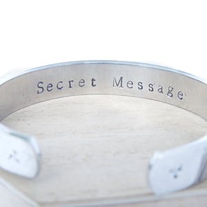 Puede incluir: Una pulsera de puño plateada con las palabras "Secret Message" estampadas en el interior. La pulsera tiene un diseño sencillo y abierto y se muestra sobre una superficie de color claro.