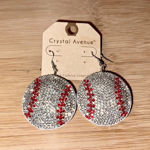 May include: A pair of baseball-themed earrings. Each earring is a round, white base covered in sparkling silver rhinestones. Red rhinestones form the baseball stitching. The earrings hang from silver fish hook ear wires. The tag reads "Crystal Avenue".
