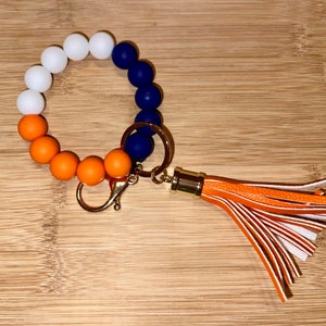 May include: A white, orange, and blue silicone beaded keychain with a gold key ring and a tassel. The tassel is orange and white.