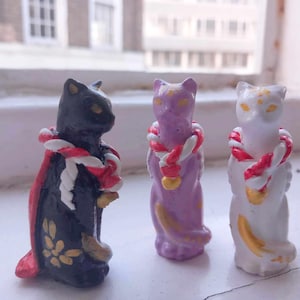 Puede incluir: Tres figuras de gato de cerámica en negro, lavanda y blanco, cada una adornada con un collar de cuerda roja y blanca. El gato negro tiene una capa roja con detalles florales dorados. Los gatos están en un alféizar.