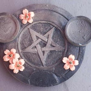 Könnte beinhalten: Ein graues, kreisförmiges Dekorationselement mit einem Pentagramm in der Mitte, zwei kreisförmigen Haltern und drei pfirsichfarbenen Blumenakzenten. Das Element hat ein strukturiertes, steinartiges Aussehen.