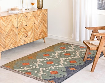 May include: A rectangular area rug with a geometric pattern in shades of green, blue, orange, and cream. The rug is placed on a light-colored floor in a room with wooden furniture and a window.