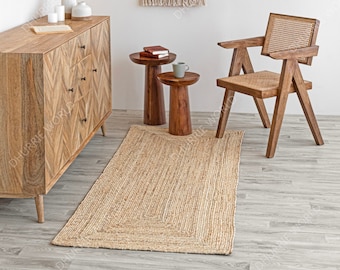 May include: A natural fiber rug in a light beige color with a woven texture. The rug has a rectangular shape with a border around the edges. The rug is placed on a wooden floor in a room with a wooden chair, two wooden side tables, and a wooden cabinet.