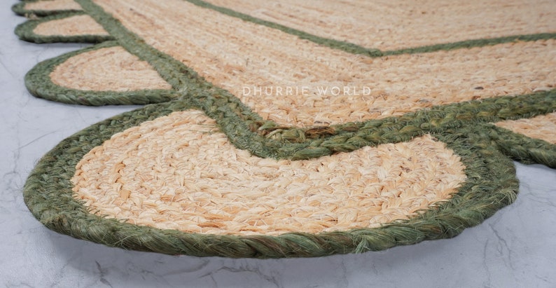 May include: A close-up of a jute rug with a scalloped edge and a green braided trim. The rug is made of natural fibers and has a rustic, earthy look.