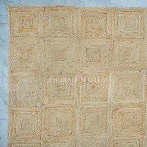 Puede incluir: Una alfombra de fibra natural con un patr&oacute;n cuadrado. La alfombra est&aacute; hecha de yute y tiene un color beige claro.