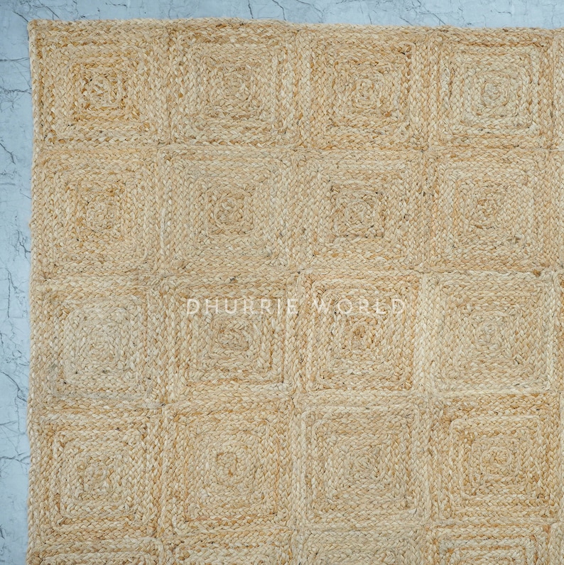 Puede incluir: Una alfombra de fibra natural con un patr&oacute;n cuadrado. La alfombra est&aacute; hecha de yute y tiene un color beige claro.