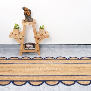 May include: A natural fiber rug with a blue scalloped border. The rug is made of jute and has a rectangular shape. The text "DHURRIE WORLD" is printed in the center of the rug.