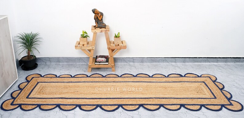 May include: A natural fiber rug with a blue scalloped border. The rug is made of jute and has a rectangular shape. The text "DHURRIE WORLD" is printed in the center of the rug.