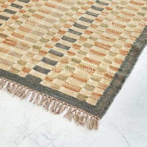 以下が含まれることがあります： 幾何学模様の長方形ラグ。ベージュ、茶色、緑、グレーの色合いの長方形が繰り返されるパターンです。濃いグレーの縁と、片側にフリンジが付いています。