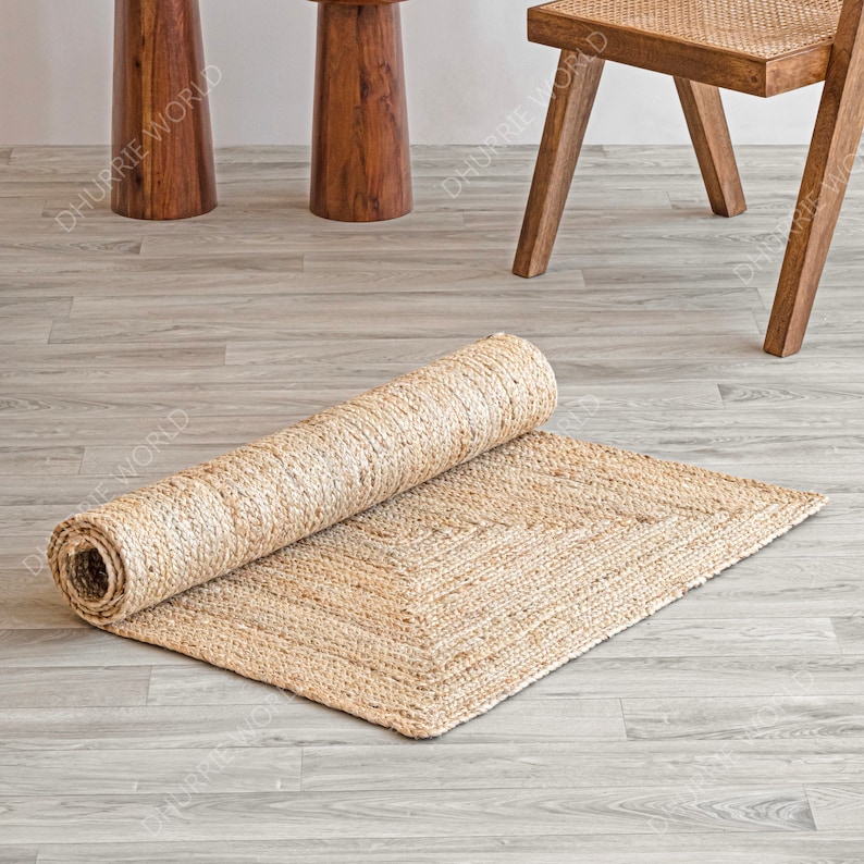 May include: A natural fiber rug made of jute, rolled up and partially unfurled on a light wood floor. The rug is a light beige color and has a woven texture.