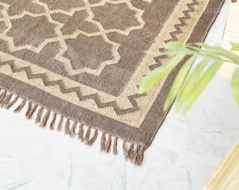 May include: A brown and beige geometric patterned rug with a fringed edge. The rug features a repeating star-like design in beige against a brown background. The border has a zig-zag pattern.